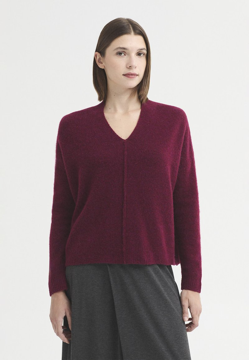 Femme aux cheveux bruns raides portant un pull bordeaux à col en V et une jupe gris foncé, debout devant un fond blanc uni.