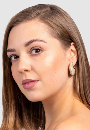 Goldene Creolen mit strukturiertem Oberflächen, besetzt mit klaren Strasssteinen, bei einem Modell mit langen, glatten Haaren auf einem grauen Hintergrund präsentiert.
