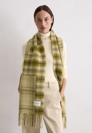 Zielony szal w kratę wykonany z miękkiej tkaniny, z frędzlami na brzegach i tkana etykietą, noszony na jasnym, prążkowanym golfie i beżowych spodniach.