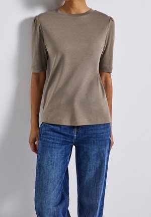 Kobieta ubrana w taupe'owy top z krótkim rękawem z bufkami na ramionach i średnio niebieskie dżinsy stojąca przy jednolitej białej ścianie.