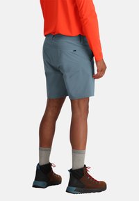 Grijze korte broek voor heren met een gladde textuur, gecombineerd met bruine wandelschoenen en grijze sokken, gezien vanuit een achteraanzicht. Helder oranje lange mouwen shirt.