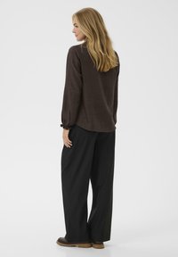 Braun strukturiertes Langarmoberteil mit elastischen Bündchen, kombiniert mit lockeren schwarzen Hosen und braunem Schuhwerk. Minimalistisches Design vor einem hellen Hintergrund.