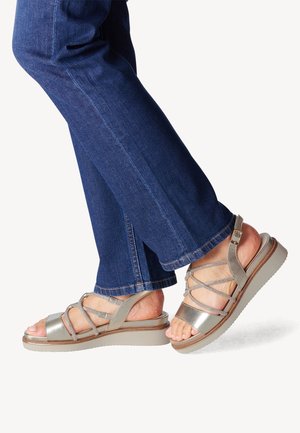 Fødder iført metalliske kilehælssandaler med stropper kombineret med blå denimjeans, den ene fod let hævet på en hvid baggrund.