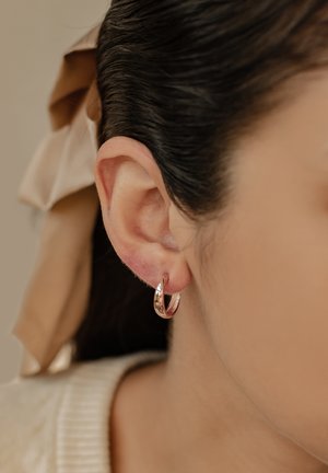 Boucles d'oreilles créoles en or rose avec une finition polie, présentant un léger design en torsion. Portées sur un lobe, elles complètent une peau lisse et des cheveux foncés.