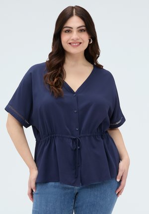 Uśmiechnięta kobieta z długimi brązowymi włosami, ubrana w granatową koszulę zapinaną na guziki z paskiem w talii oraz jasnoniebieskie dżinsy na jednolitym jasnym tle.