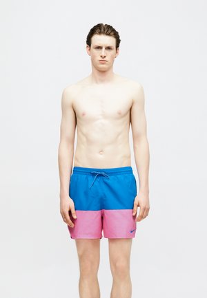 Junger, oberkörperfreier Mann steht frontal und trägt blau-pinke, farblich abgestufte Badeshorts mit Kordelzugbund.