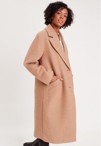 Beige pitkä villatakki, jossa on läpikuultava kaulus, etupocketit ja rento istuvuus. Sujuva pinta, jossa on hienovaraiset ompeluyksityiskohdat.