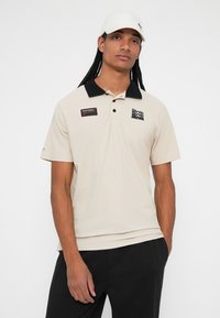Beige polo skjorte med sort krave. Har et logo og et mærke på brystet. Glat tekstur, korte ærmer. Bæres med mørke bukser og en hvid kasket.