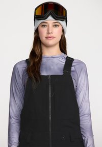 Combinaison de ski noire avec fermeture éclair à l'avant, portée sur un t-shirt à manches longues violet clair. Un bonnet en tricot gris et des lunettes de ski à visière sombre complètent la tenue.