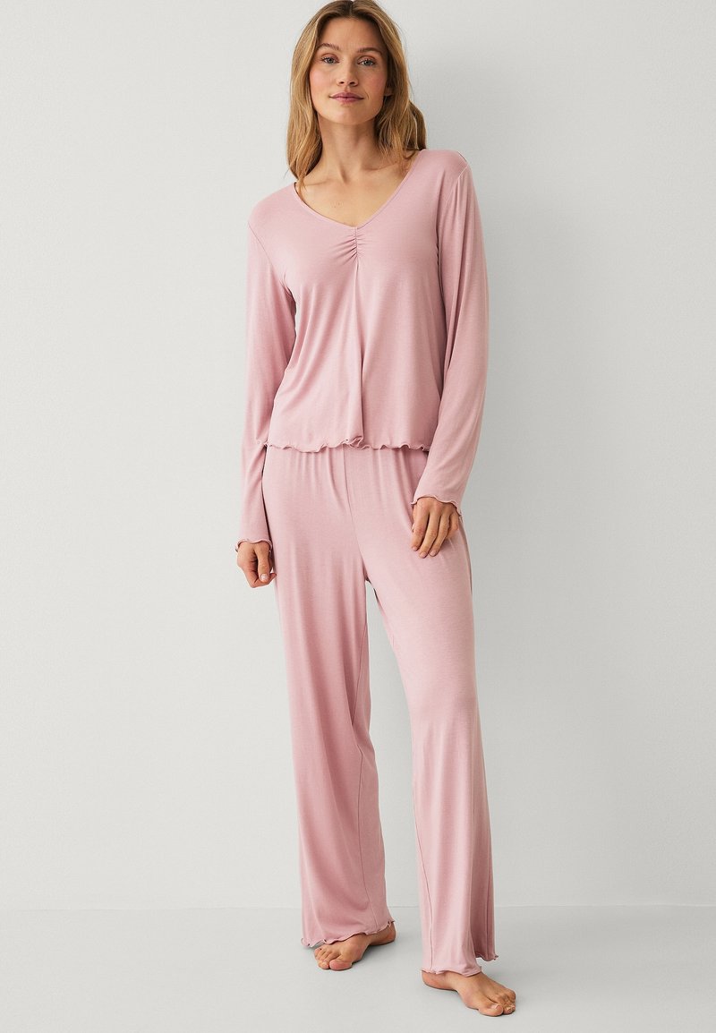 Kobieta stojąca boso, ubrana w miękki, różowy top z długim rękawem z falbanką na dole oraz dopasowane, luźne spodnie na jednolitym jasnym tle.