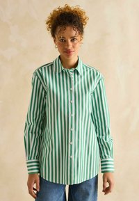 Grün-weiße gestreifte Button-Down-Bluse mit langen Ärmeln, klassischem Kragen und abgerundetem Saum. Getragen mit blauen Jeanshosen.