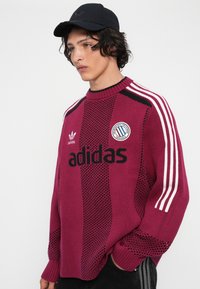 Jauns vīrietis, ģērbies maroon Adidas džemperī ar baltām svītrām uz piedurknēm un melnā cepurē, stāvot ar vienu roku kabatā.