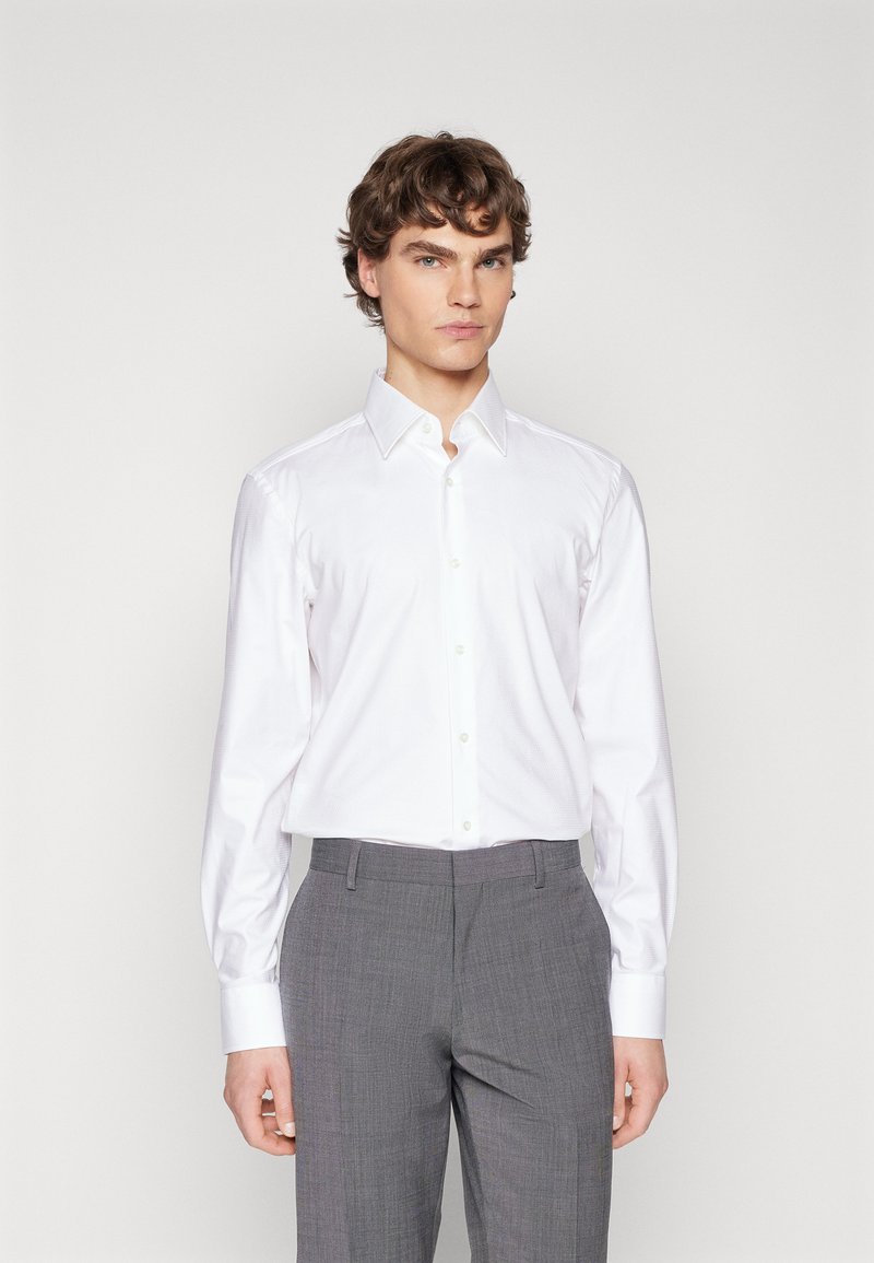 Camicetta bianca a maniche lunghe con colletto a punta, abbinata a pantaloni grigi con texture, caratterizzati da una vestibilità slim e tessuto morbido.