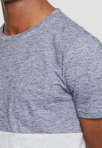 Hombre que lleva una camiseta de manga corta en tono gris azulado jaspeado con un bolsillo en el pecho y una sección inferior de un gris más claro.