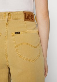 Vue arrière d'une personne portant un jean Lee marron clair avec une poche arrière visible et un patch en cuir de la marque Lee sur la ceinture.