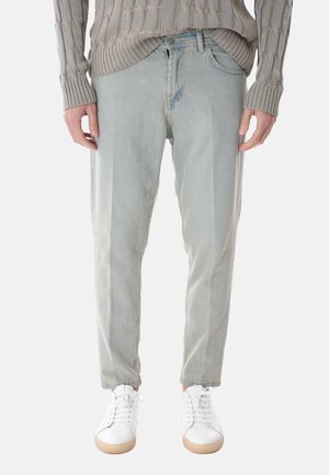 Persona che indossa jeans grigi chiari a gamba dritta, maglione beige lavorato a trecce e scarpe da ginnastica bianche con suole marrone chiaro, in piedi contro uno sfondo neutro.