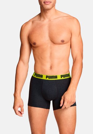 Schwarze Boxer-Shorts mit einem gelben Logo am Bund, mit einer körperbetonten Passform und glatter Textur, präsentiert von einer männlichen Figur mit neutralem Gesichtsausdruck.
