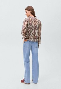 Blouse translucide à motifs dans des nuances de crème, bordeaux et marron, associée à un jean droit bleu clair et des baskets rouges.
