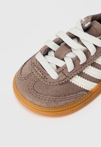 Sapatilhas de camurça castanha com atacadores brancos, riscas laterais brancas e uma sola translúcida de borracha texturizada, mostradas de um ângulo próximo e superior.