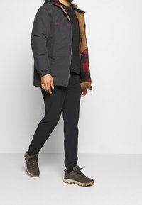 Uomo che cammina indossando pantaloni neri, sneakers verde scuro e marroni, maglione nero e giacca invernale nera con cappuccio foderato in pelliccia e rivestimento a scacchi rosso.