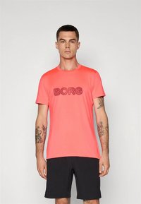 Koralen jopič z napisom "BORG" v kontrastnih črkah, kratkimi rokavi in okroglim izrezom, kombiniran s črnimi kratkimi hlačami.