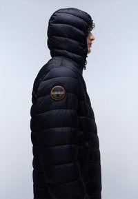 La giacca a vento nera presenta un design trapuntato, un cappuccio e una cerniera laterale. Include una patch circolare con un logo sul braccio sinistro.