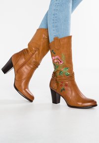 Braune Lederstiefeletten mit Absatz, die mit floralen Stickereien, einem Reißverschluss und plissierten Details versehen sind. Das Design umfasst einen Blockabsatz und Quastenakzente.