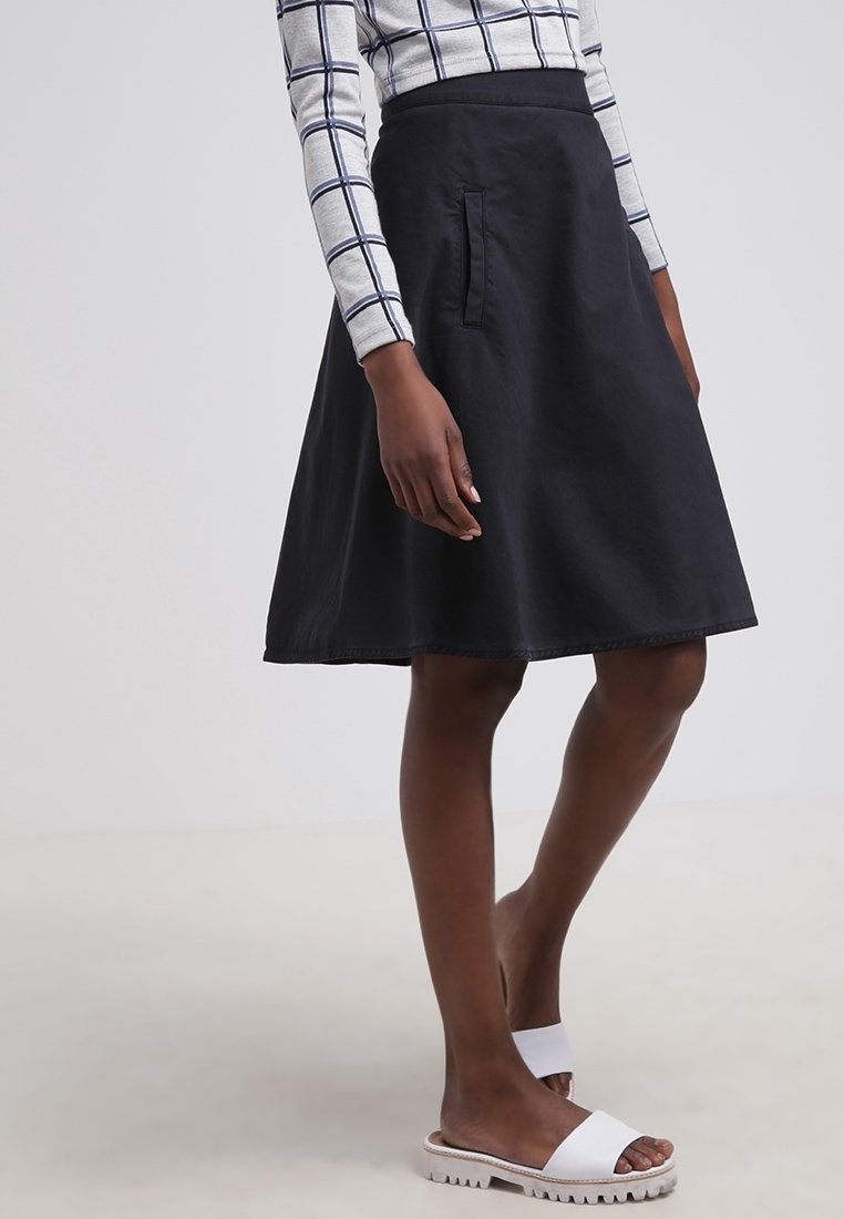 Jupe noire en forme de A avec des poches latérales et une texture lisse. Associée à un haut à manches longues à carreaux et des sandales blanches.