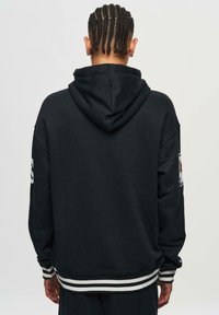 Black hooded sweatshirt featuring a relaxed fit, ribbed cuffs and hem with white stripes, and a branded patch on the left sleeve.