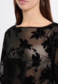 Schwarze transparente Bluse mit floralem Samt-Applikationen, runder Halslinie und langen Ärmeln, die ein zartes und strukturiertes Design zeigt.