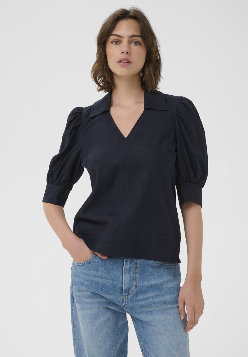 Femme debout, une main dans la poche, portant un chemisier noir à col en V avec des manches bouffantes et un jean bleu clair, fond gris neutre.