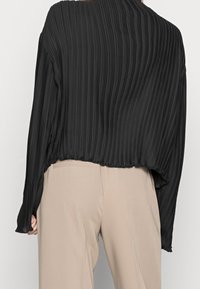 Top lungo a maniche lunghe plissettato nero con una vestibilità rilassata e orlo arrotondato. Il tessuto sembra leggero con una texture liscia. Abbinato a pantaloni beige.