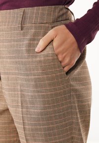 Pantaloni a quadri marroni con tessuto testurizzato, tasche laterali e vestibilità su misura. Una mano è appoggiata nella tasca destra.