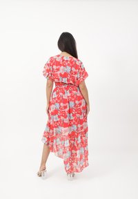 Robe maxi fleurie rouge avec des manches courtes, taille cintrée et ourlet à volants. Présente des fleurs bleu clair et blanches sur un tissu transparent.