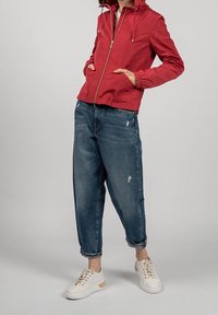 Rød letvægtsjakke med lynlåsfortil, elastiske manchetter og justerbar hætte. Båret over slidte blå denimbukser og hvide sneakers.