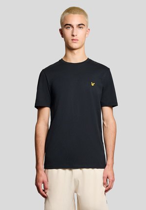 Ung mand med kort blondt hår iført en sort t-shirt med en lille gul fuglelogo og lyse beige bukser mod en grå baggrund.
