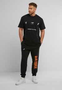 Crna pamučna majica s natpisom "Sveučilište Floride", u kombinaciji s crnim trenerkama koje sadrže narančasti natpis "GATORS". Bijele tenisice upotpunjuju izgleda.