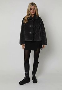 Veste en fausse fourrure gris foncé avec trois fermetures à bascule, design oversize et manches larges, associée à une mini-jupe noire et des bottes montantes jusqu'aux genoux.