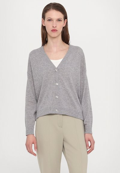 Pelēks kardigans ar V veida kakla izgriezumu, piecām pērļu pogām un nokareniem pleciem. Mīksta tekstūra un īsā garumā; kombinēts ar gaiši bežām biksēm.