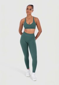 Reggiseno sportivo verde e leggings abbinati, realizzati in materiale liscio ed elastico. Scarpe da ginnastica bianche e design minimalista senza motivi.