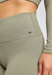Leggings côtelés vert olive avec une taille haute, arborant une étiquette logo métallique et une texture lisse. La vue en gros plan met en évidence les détails de couture.