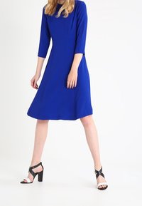 Niebieska sukienka z okrągłym dekoltem, rękawami o długości 3/4 i rozkloszowanym dołem, w zestawieniu z czarno-białymi sandałami na wysokim obcasie.