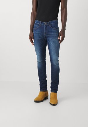 Osoba nosząca obcisłe niebieskie dżinsy i musztardowo-żółte zamszowe botki, stojąca na jednolitym jasnym tle.