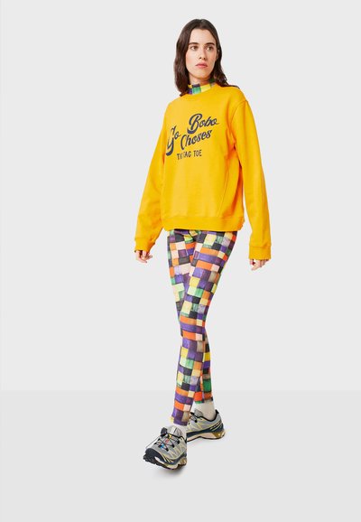 Felpa gialla con testo grafico abbinata a leggings colorati e a fantasia che presentano quadrati in viola, arancione, verde e beige.