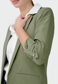 Blazer vert olive en tissu lisse, doté d'un col cranté, de manches retroussées avec détail boutons, et d'une poche avant.