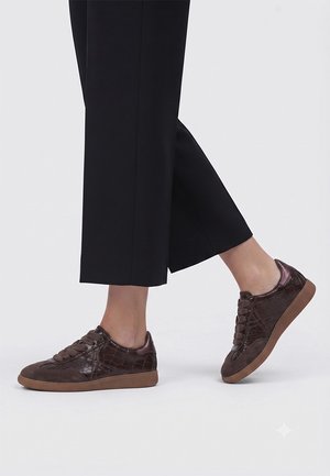 Persona que lleva pantalones cortos negros y zapatillas texturizadas marrón oscuro con suelas de goma, mostrada desde las pantorrillas hacia abajo sobre un fondo liso.