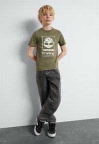Groene katoenen t-shirt met witte boomegrafiek en "TIMBERLAND" tekst. Grijze baggy jeans en zwarte sneakers met witte zolen.