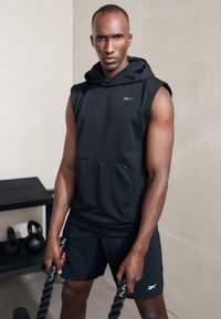 Fekete ujjatlan kapucnis pulóver elülső zsebből és kis logóból, összehangolt sorttal párosítva; a kezek fekete kötelet szorítanak egy zárt térben.