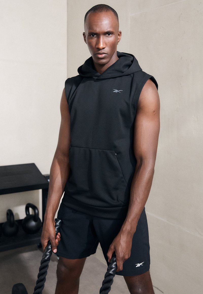 Fekete ujjatlan kapucnis pulóver elülső zsebből és kis logóból, összehangolt sorttal párosítva; a kezek fekete kötelet szorítanak egy zárt térben.