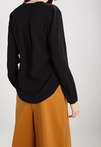 Schwarze Bluse mit langen Ärmeln und rundem Ausschnitt, Rückenschlitzdetail und leicht abgerundetem Saum, kombiniert mit hochtaillierten senfgelben Hosen.